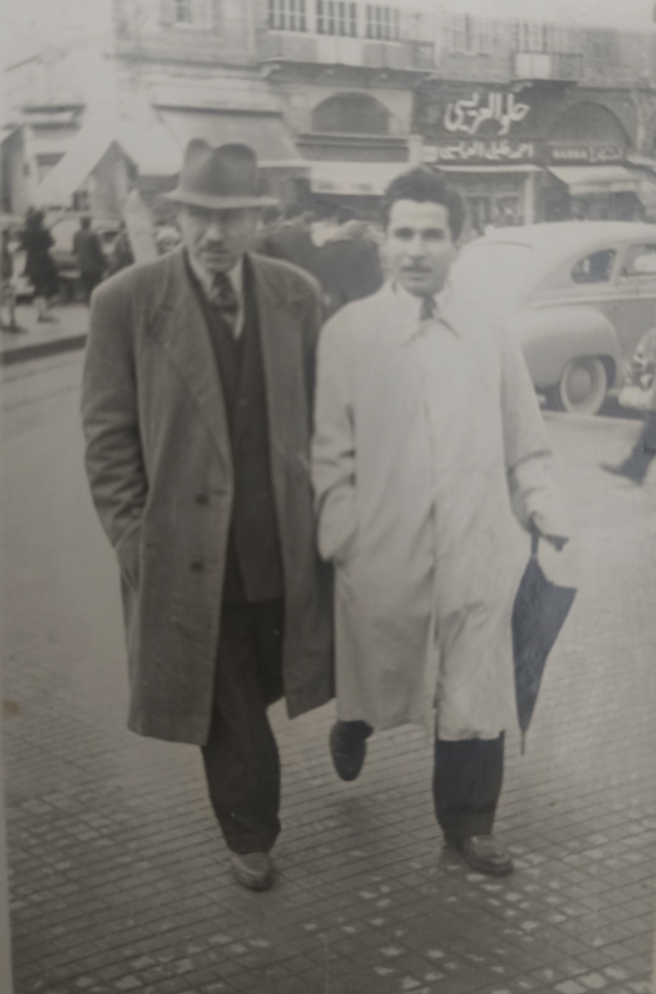 Bir caddede yürüyen iki kişi, arkada Arapça tabelalı dükkânlar — Mustafa İloğlu arşivi